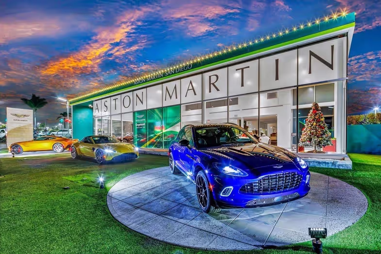 Blue Aston Martin displayed beside a decorated dealership showroom at dusk