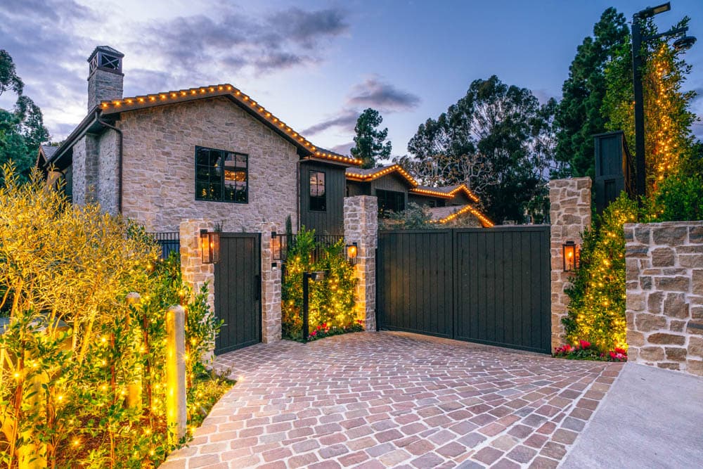 Stone estate entry, rooflines, and landscape illuminated with warm holiday lights in an HLI portfolio image